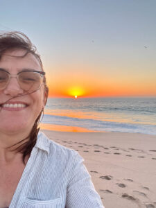 A selfie of a woman with the sunrise behind her, over a beach. Her hair is windblown and she is smiling.